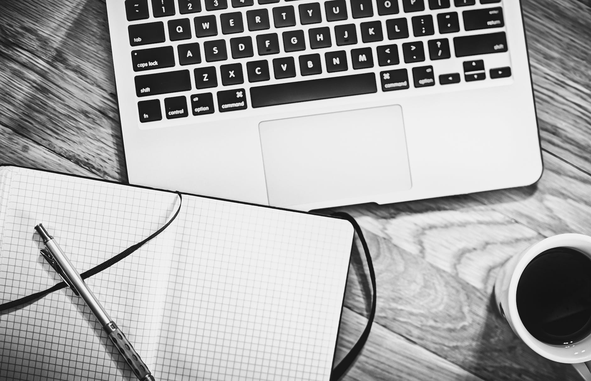um caderno aberto, caneta, teclado de um laptop e uma xícara de café