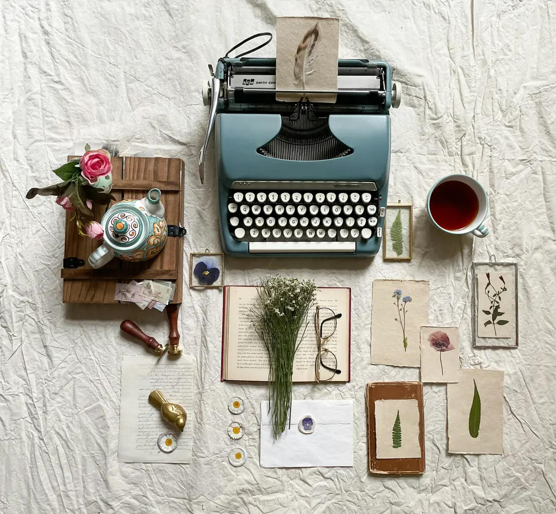 fotografia de uma máquina de escrever, cercada por itens diversos como óculos, papéis, flores, xícara, etc